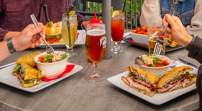Several plates of food on a patio table including sandwiches, salads, and beer.
