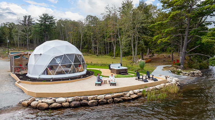 Geodesic dome (yurt) glamping accommodation beside a lake with seating area and hot tub.