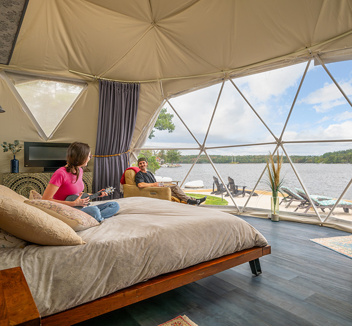 Interior of a riverside geodesic dome with a bed and large window overlooking the water.