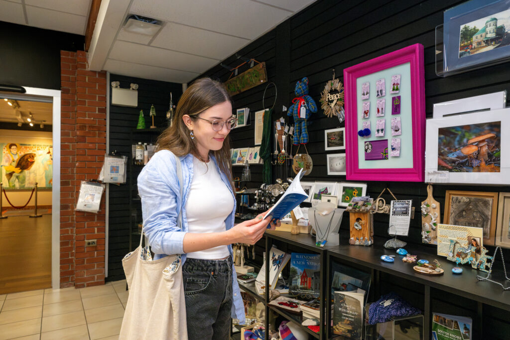 A woman stands next to a display of gift shop items at the DesBrisay museum. She holds a book in her hands. Behind her, the display shows a colourful array of artwork, jewellery, and books for sale.