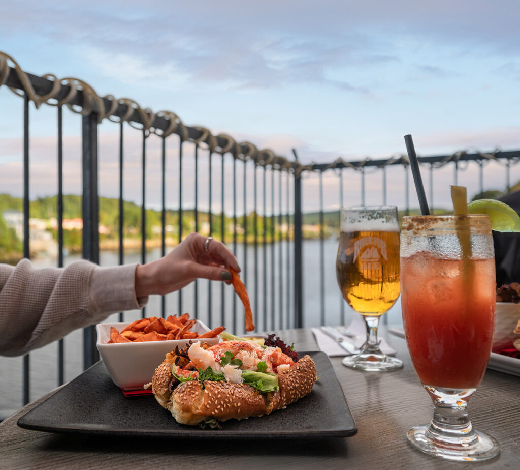 Close-up shot of a hand holding a french fry, with a bread roll filled with lobster and two beverages.