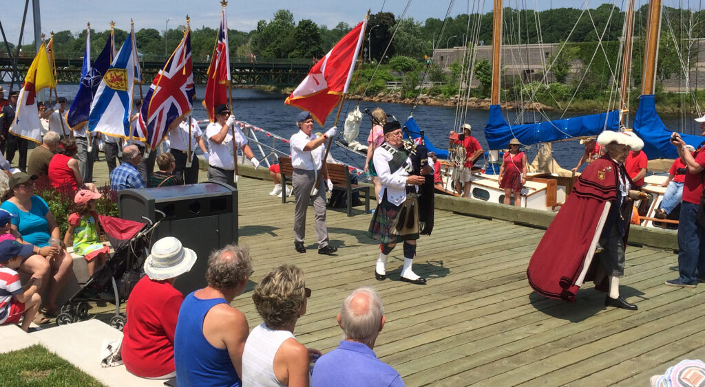 A crowd on the LaHave River waterfront watches as a parade goes by. A man playing bagpipes can be seen leading several people whom are carrying various flags.