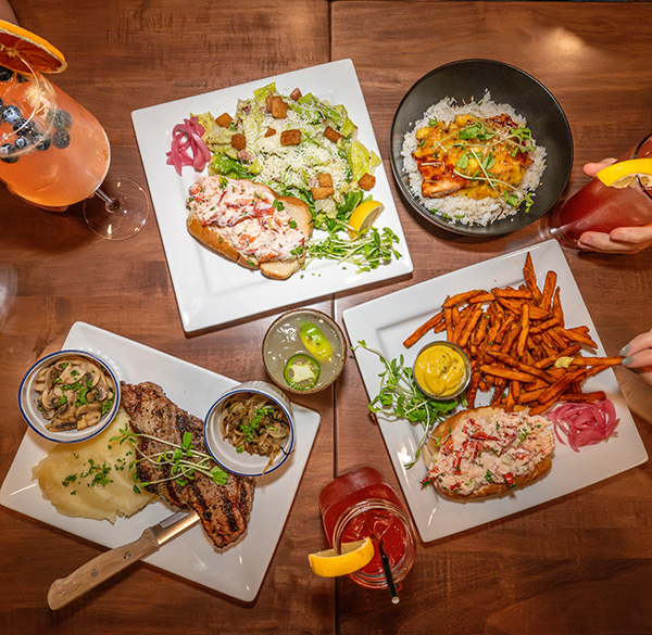 Restaurant table with lobster roll, grilled steak, sweet potato fries, salad, and cocktails.